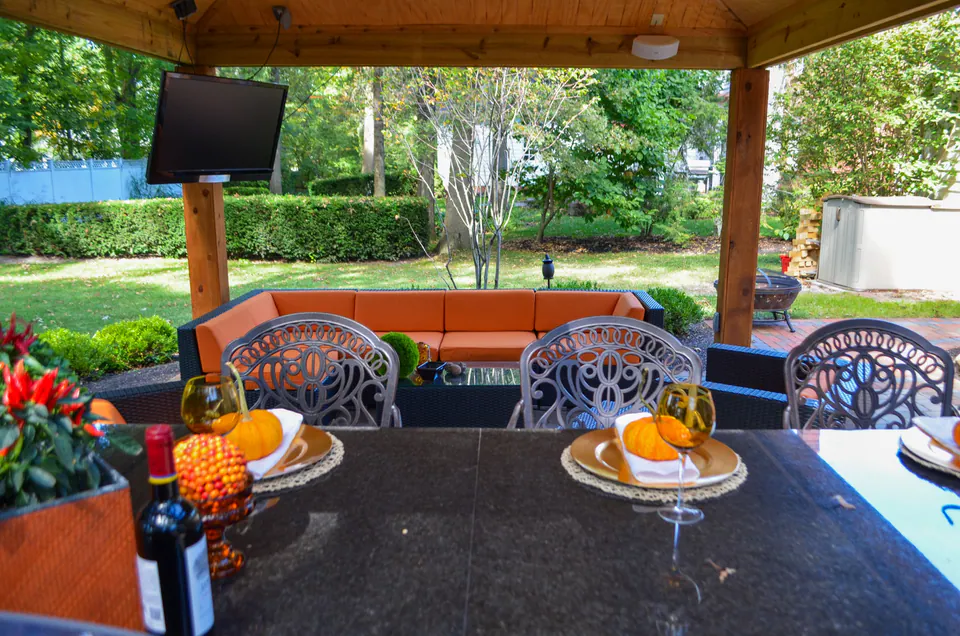 Dining table with pumpkins under covered pavilion, orange bench seating, television, wood posts, and backyard view.